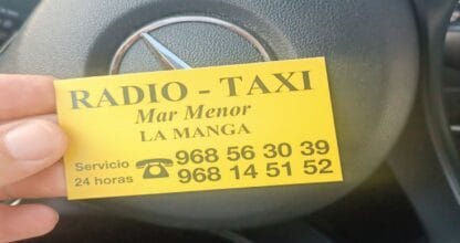 Tarjeta amarilla de Radio-Taxi Mar Menor La Manga con servicio 24 horas sostenida frente al volante